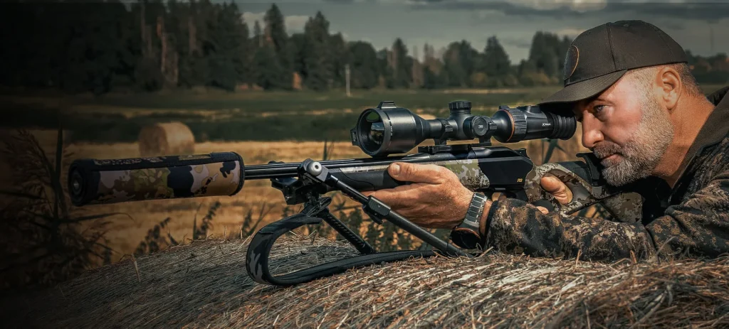 Hunter aiming a ACE thermal imaging riflescope while resting on a hay bale.