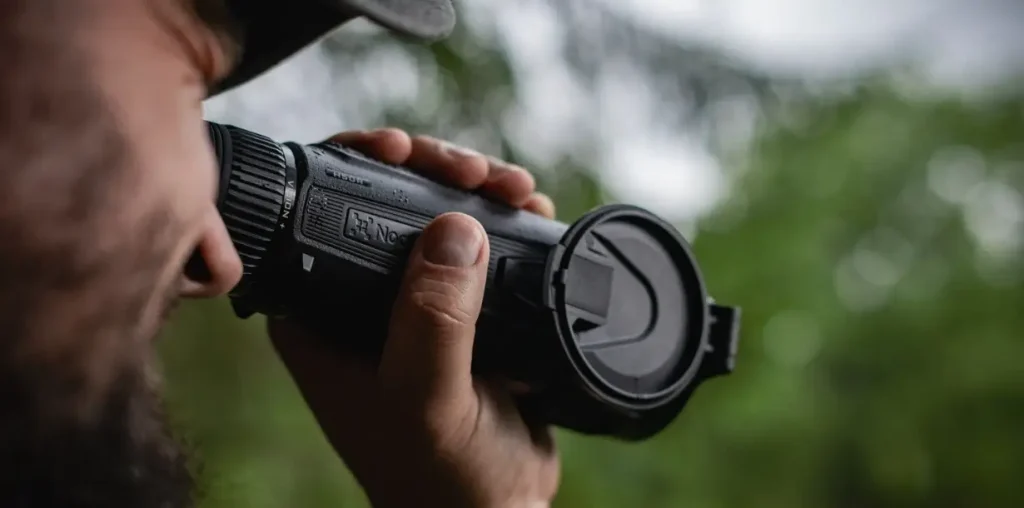 Man viewing through Nocpix H50R thermal monocular with open lens cover outdoors.