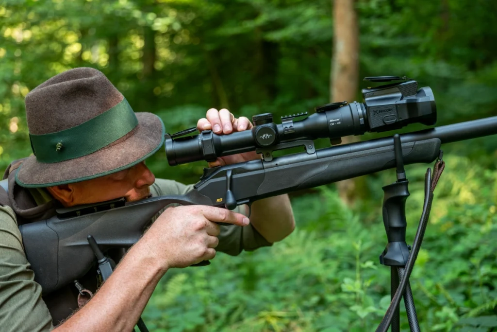 Hunter aiming a rifle equipped with a thermal scope while positioned.