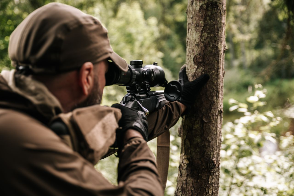 Hunter aiming a scoped rifle while using a tree for support in a forest.