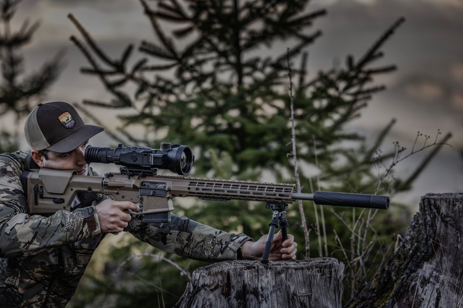 Unter Zielen mit einem schallgedämpften Gewehr mit Wärmebildzielfernrohr, das auf einem Zweibein in einem Waldgebiet gestützt wird.