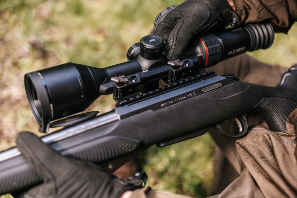 A person adjusting the Nocpix ACE RM thermal imaging riflescope mounted on a bolt-action rifle outdoors.
