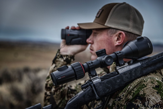 Hunter using a rangefinder while carrying a rifle with mounted scope during a hunting scouting session.