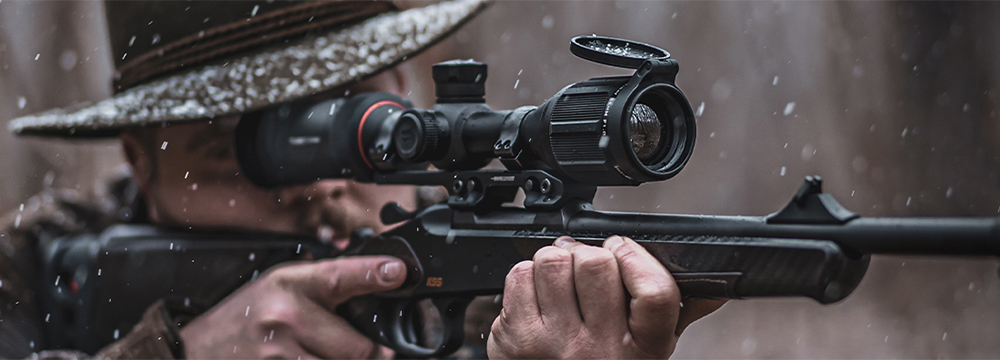 Person wearing a hat aiming a rifle with a mounted scope in rainy outdoor conditions.