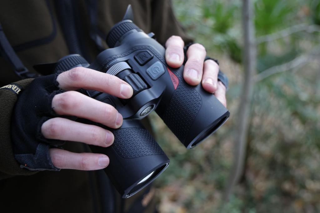 Hands holding black binoculars outdoors in a wooded setting