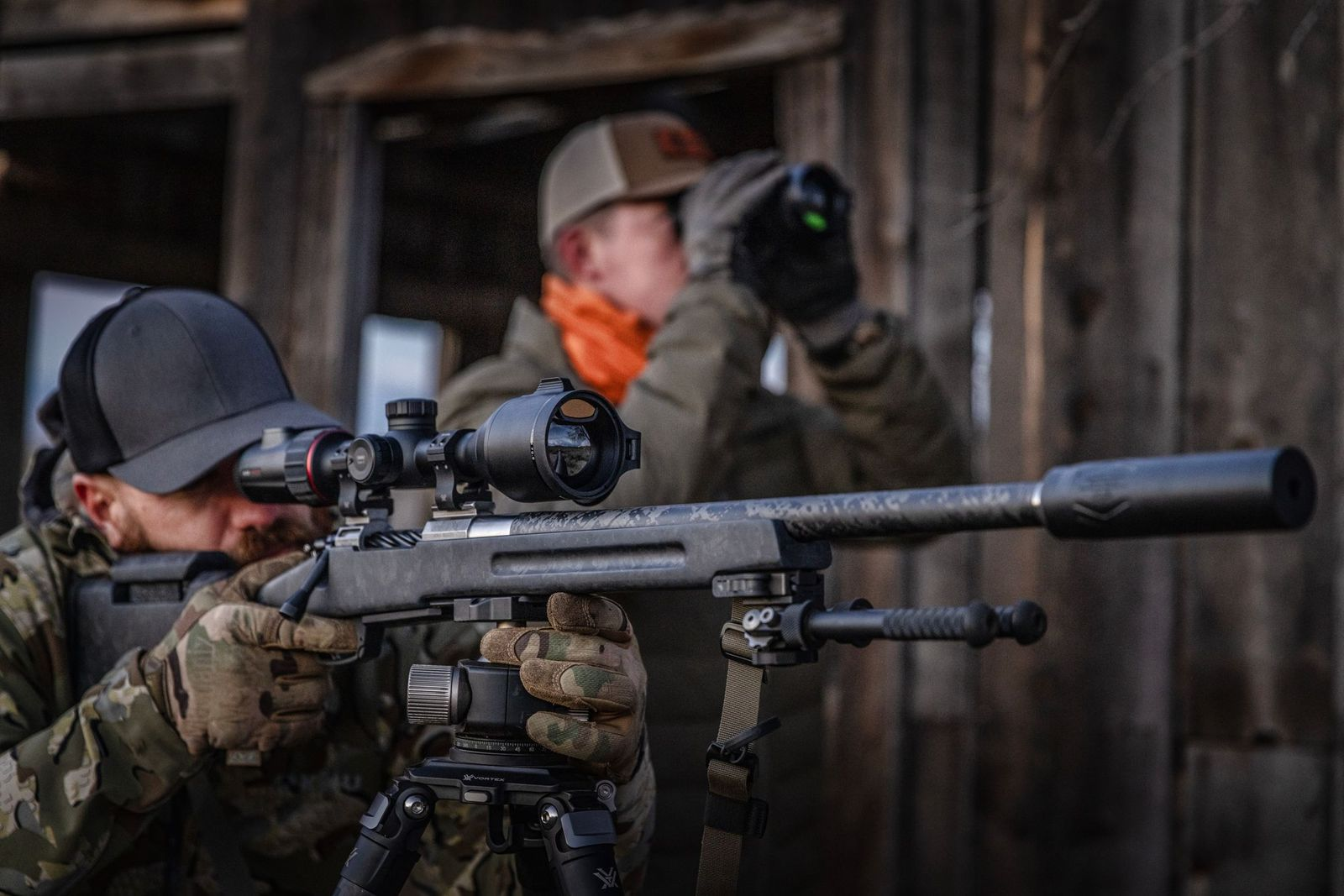 Shooter aiming a scoped rifle on a tripod while a spotter uses binoculars in the background