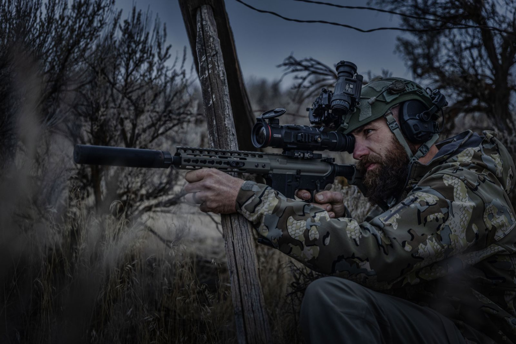 Person in camouflage gear aiming a rifle with an optical sight in an outdoor wooded environment.