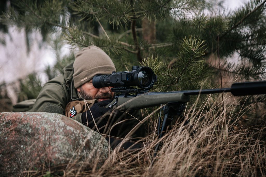 Hunter aiming with a scoped rifle from cover in grassy terrain