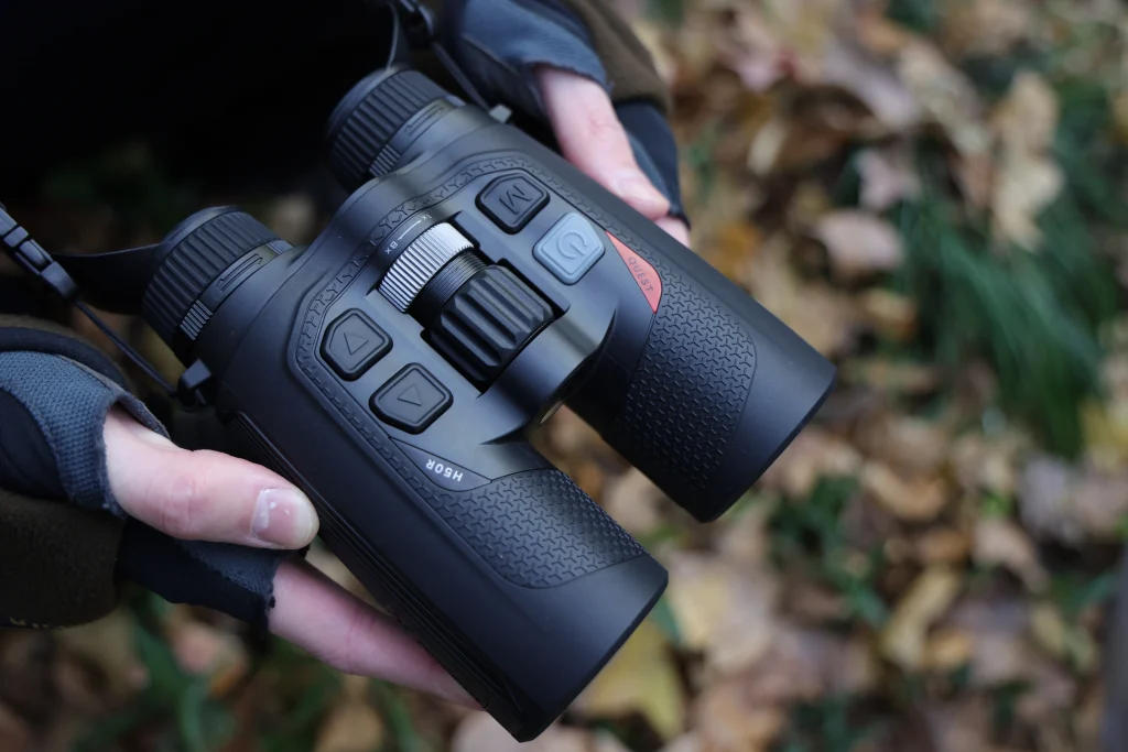 A hunter holds black thermal binoculars while scouting in woodland terrain.
