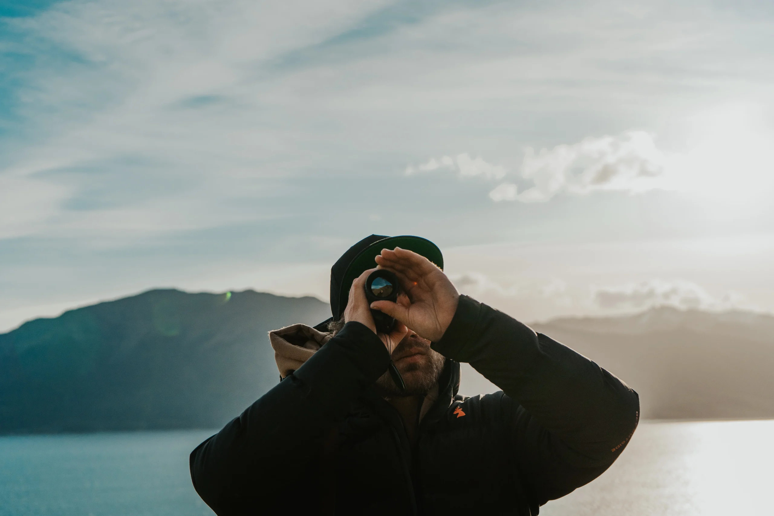 A hunter looks through a handheld thermal monocular near a lake and mountain landscape.